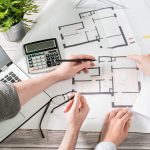 two people sit at a desk with a laptop and calculator working on the floor plan design for a new home.