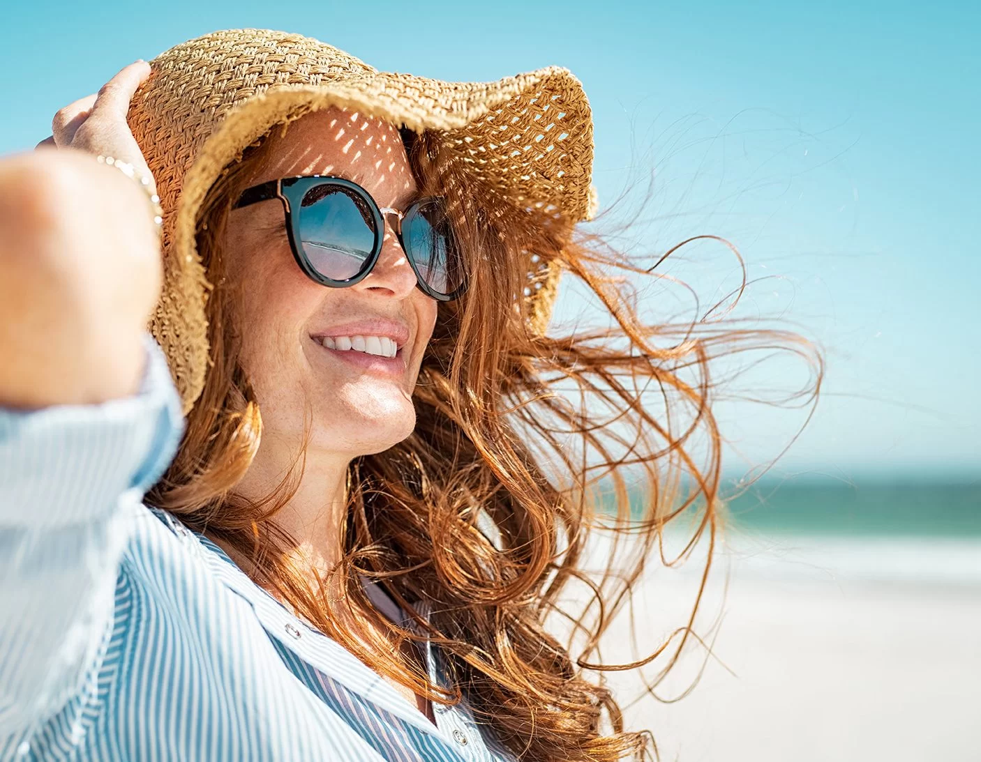Smiling woman holding her sun hat in the breeze by the water