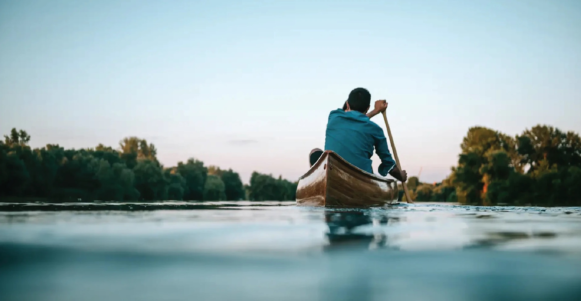 Man paddling a wooden canoe on a calm river at dusk