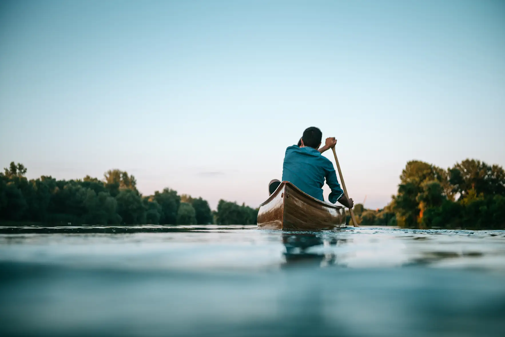 Man paddling a wooden canoe on a calm river at dusk